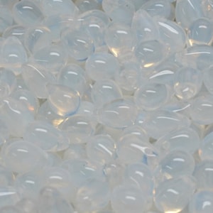 May include: A close-up shot of a collection of opalescent, teardrop-shaped beads. The beads are a milky white color with a subtle, pearlescent sheen. They appear smooth and rounded, suggesting a polished finish. The beads are scattered, filling the frame.