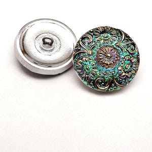 Op de afbeelding: Knop in twee delen met een zilveren basis en een decoratieve bovenkant. De bovenkant heeft een sierlijk bloemmotief met iriserende tinten teal, paars en brons. De knop is waarschijnlijk voor naaien of knutselen.