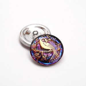 Op de afbeelding: Twee ronde knopen met een metallic zilveren achterkant en een oogje. De voorste knoop toont een goudkleurige vogel op een tak tegen een donkere, iriserende paarse en blauwe achtergrond met een glanzende afwerking en reliëf.