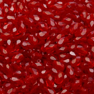 May include: A close-up view of a large quantity of small, faceted, red glass beads. The beads are oval shaped and have a glossy finish.