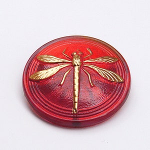 Puede incluir: Un botón de vidrio rojo con un diseño de libélula dorada. El botón tiene una superficie texturizada en relieve.