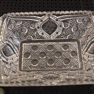 May include: A clear glass square dish with intricate cut-glass patterns. The dish has a raised center section with a smaller square pattern.