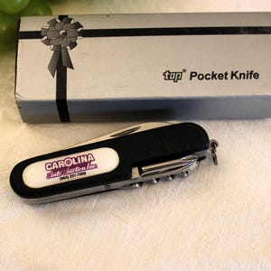 May include: A black pocket knife with a white label that says "CAROLINA State Station Inc. (843) 231-7000". The knife is in a silver box with a black ribbon and the text "top Pocket Knife".