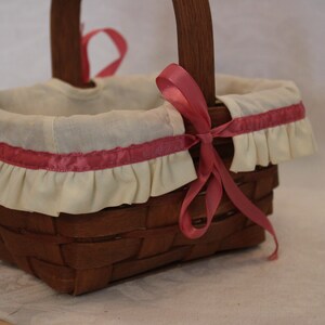 May include: A small, brown wicker basket with a white fabric lining and a pink ribbon. The basket has a ruffled edge and is decorated with a pink ribbon.