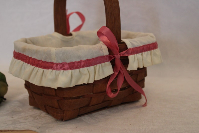 May include: A small, brown wicker basket with a white fabric lining and a pink ribbon. The basket has a ruffled edge and is decorated with a pink ribbon.