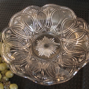 May include: A clear glass bowl with a scalloped edge and intricate cut-glass design. The bowl has a starburst pattern in the center and a repeating pattern of arches around the edge.
