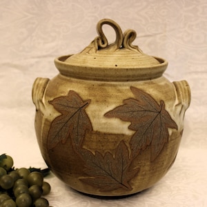 May include: A ceramic jar with a lid, decorated with brown leaves. The jar has two handles on the sides and a unique, twisted handle on the lid.