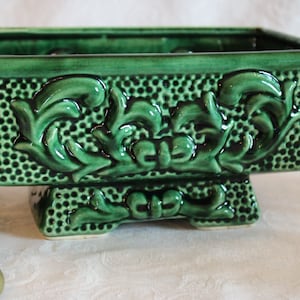 Peut inclure: Un pot en céramique vert avec un motif floral en relief. Le pot a une base carrée avec quatre pieds.