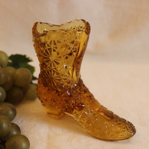 May include: A small amber glass boot-shaped vase with a diamond pattern. The vase is sitting on a white surface with a bunch of green grapes in the background.