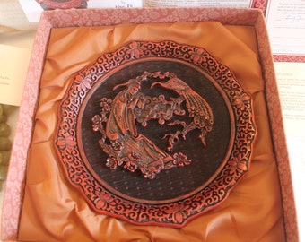 May include: A red wooden box with a silk lining and a carved wooden plate depicting a woman and a bird. The plate is surrounded by a decorative border.