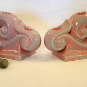 May include: A pair of pink and green ceramic candle holders with a swirl design. The holders are shaped like a seashell and have a square base.