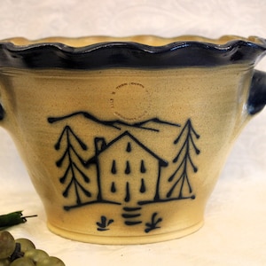 May include: A yellow ceramic bowl with blue trim and a black silhouette of a house with two trees on either side. The bowl has two handles and a scalloped rim. The bowl is stamped with "The Old Mill Pottery, Somerset, Ohio".