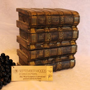 May include: A stack of wooden book sculptures with brass accents. The books are painted in shades of brown and gold, with a weathered appearance. A bunch of grapes and a tag that reads "SEPTEMBER WOODS" are in the foreground.