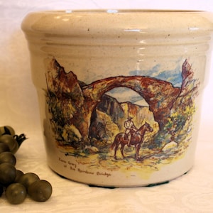 May include: A beige ceramic crock with a painted image of a natural stone arch bridge. The image also includes a person on horseback riding through the arch. The text on the crock reads "Rainbow Bridge, Natural Bridges National Monument, Utah".
