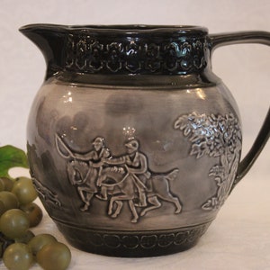 May include: Gray ceramic pitcher with a dark gray rim and handle. The pitcher features a relief design of figures on horseback and a tree. A bunch of green grapes rests in the foreground.