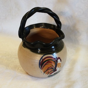 May include: A ceramic rooster-themed pitcher with a black handle. The pitcher has a scalloped rim and is decorated with a rooster in shades of red, orange, yellow, and green.