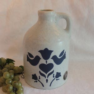 May include: A white ceramic pitcher with a blue and white floral design. The pitcher has a handle and a spout. The design features two birds and a heart.
