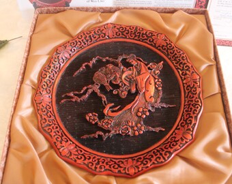 May include: A red and black lacquered wooden plate with a carved design of a woman standing in a tree with birds. The plate is in a box with a brown satin lining.