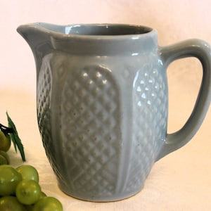 May include: A small, light blue ceramic creamer with a textured, diamond-shaped pattern. The creamer has a handle and a spout.