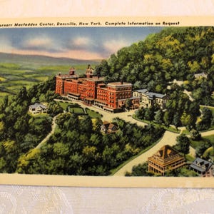 May include: A vintage postcard depicting a large, multi-story building with a green roof and white trim. The building is surrounded by trees and is set against a backdrop of rolling hills. The text on the postcard reads "Bernarr Macfadden Center, Dansville, New York. Complete Information on Request."