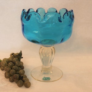 May include: A blue glass bowl with a ruffled rim sits on a clear glass pedestal. The bowl is on a white surface with a bunch of green grapes.