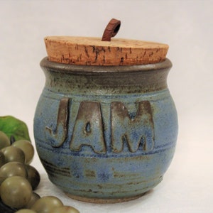 May include: A handmade ceramic jam jar with a cork lid. The jar is blue with brown lettering that spells "JAM". The lid has a leather pull. The jar is a rustic, country-style kitchen item.