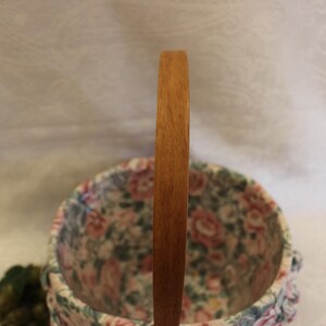 May include: A floral patterned fabric basket with a wooden handle. The basket is round and has a white background with pink, red, and green flowers.