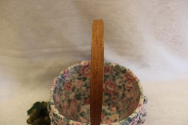 May include: A floral patterned fabric basket with a wooden handle. The basket is round and has a white background with pink, red, and green flowers.