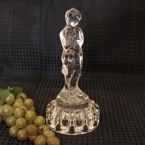 May include: A clear glass figurine of a person standing on a pedestal. The figurine is detailed and has a textured base.