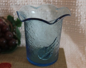 Puede incluir: Un jarrón de vidrio azul claro con una superficie texturizada y esmerilada y un borde rizado. El jarrón tiene un diseño único con un patrón circular en relieve.