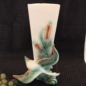 May include: A white ceramic vase with a green and brown bird design. The vase is tall and slender with a wide opening at the top. The bird is in flight and has its wings spread out. The vase is decorated with a floral design of green leaves and brown reeds.