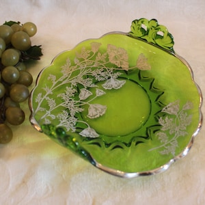 May include: A green glass serving dish with silver floral accents and two handles. The dish has a scalloped edge and is perfect for serving snacks or desserts.