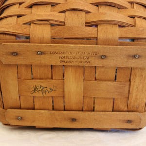 May include: A close-up of the bottom of a brown woven basket. The basket is labeled "Longaberger Basket" and "Handwoven,  Ohio, USA". The basket is signed "K.E.B. 1993".