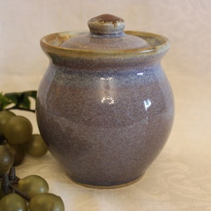 May include: A small, round ceramic jar with a lid. The jar is a light purple colour with a brown glaze around the top. The jar is sitting on a white surface.
