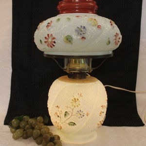 May include: A white glass lamp with a floral design. The lamp has a red top and a white cord. The base of the lamp is round and has a floral design. The lamp is sitting on a black surface.