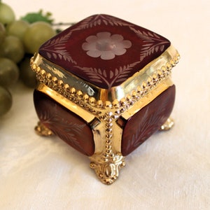 May include: A small, square, red glass trinket box with gold trim and feet. The box has a floral design etched into the glass.