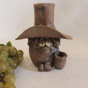 May include: A brown ceramic figurine of a person wearing a tall hat and holding a small brown pot. The figurine has a whimsical, cartoonish style.