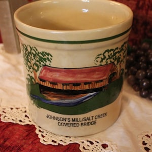 May include: A white ceramic mug with a green, brown, and red painted design of a covered bridge. The text "Johnson's Mill/Salt Creek Covered Bridge" is printed on the bottom of the mug.