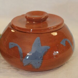 May include: A small, round, terracotta-colored ceramic jar with a lid. The jar has a blue and white abstract design painted on the side.