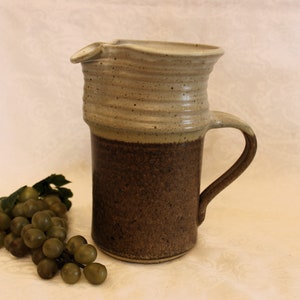 Peut inclure: Un pichet en céramique brun et blanc avec une anse et un bec verseur. Le pichet a un glaçage moucheté et une bande brune autour du milieu.