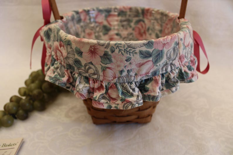 May include: A pink and green floral patterned basket with a ruffled edge and red ribbon handles.