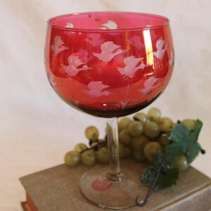 May include: A red glass wine goblet with a white etched design of flying birds. The goblet is sitting on a brown book with a bunch of green grapes in the background.