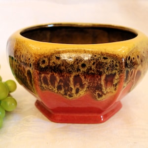May include: A red, yellow, and brown glazed ceramic planter with a hexagonal base. The planter has a mottled glaze with a drip effect.