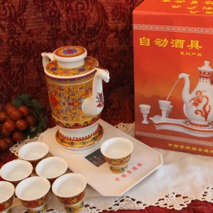 May include: A white ceramic automatic liquor dispenser set with a colourful teapot, eight small cups, and a tray. The box reads "自动酒具" which translates to "Automatic Liquor Dispenser" in English.