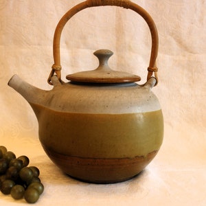 May include: A ceramic teapot with a light brown and gray glaze. The teapot has a bamboo handle and a lid. The teapot is sitting on a white surface.