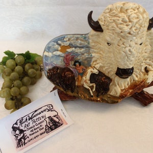 May include: A ceramic sculpture of a white buffalo with brown accents. The buffalo is depicted in a scene with a Native American figure and other buffalo. The sculpture is painted with a blue sky and clouds. The sculpture is signed "Rich Wisecarver's Art Pottery".