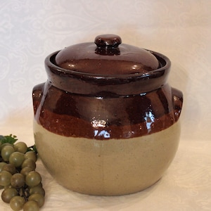May include: A brown and beige glazed ceramic crock with a lid. The crock has two handles on the sides. A bunch of green grapes are in the foreground.