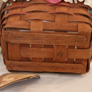 May include: A brown wicker basket with a white ruffled fabric lining and a pink ribbon. The basket has a label on the bottom that reads "Longaberger Basket Co. Handwoven in U.S.A."