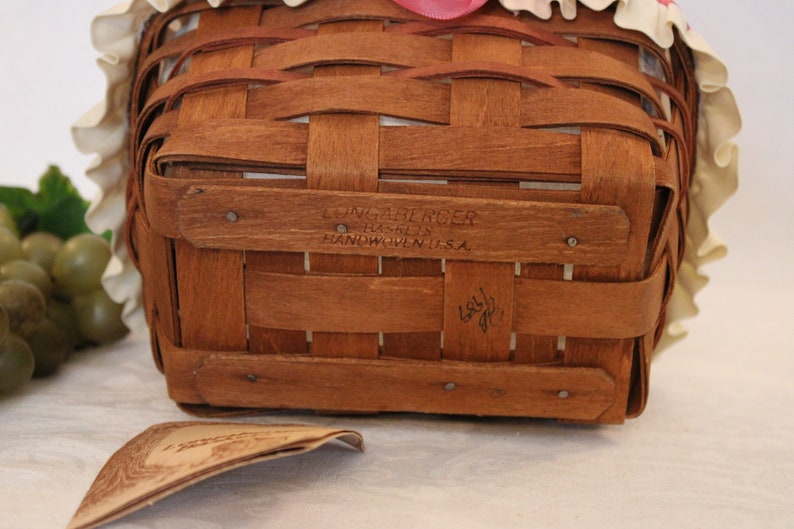 May include: A brown wicker basket with a white ruffled fabric lining and a pink ribbon. The basket has a label on the bottom that reads "Longaberger Basket Co. Handwoven in U.S.A."