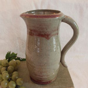 May include: A handmade ceramic pitcher with a light brown glaze and a red rim. The pitcher has a rounded body and a curved handle.